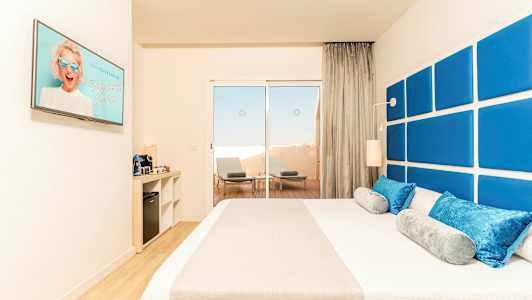 Hotellihuone, jossa sänky ja sinisiä sekä harmaita tyynyjä, sinisillä paneeleilla koristellun seinän edessä. Nurkassa on kaappi, jossa kahvikone ja minibaarijääkaappi. Suuri ikkuna johtaa terassille, jossa on kaksi aurinkotuolia ja pöytä. (Tekoälyn luoma teksti)
