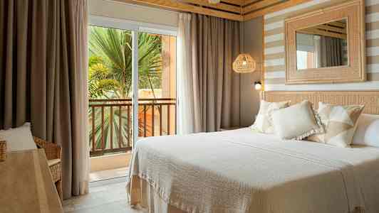 Hotellihuone, jossa on parivuode, vaaleita tekstiilejä ja sisustuselementtejä. Ulkona näkyy parveke, jolta on näkymä vehreyteen. (Tekoälyn luoma teksti)