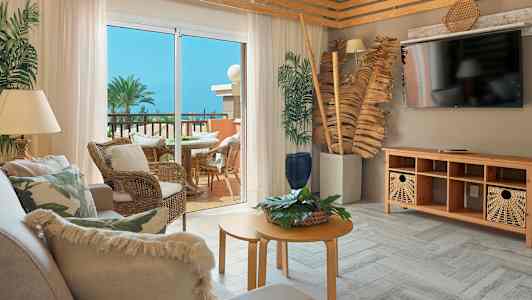 Olohuone, jossa puiset yksityiskohdat ja sohva, uloskäynti kalustetulle parvekkeelle, josta on näkymä palmuihin ja kukoistavaan kasvillisuuteen. (Tekoälyn luoma teksti)