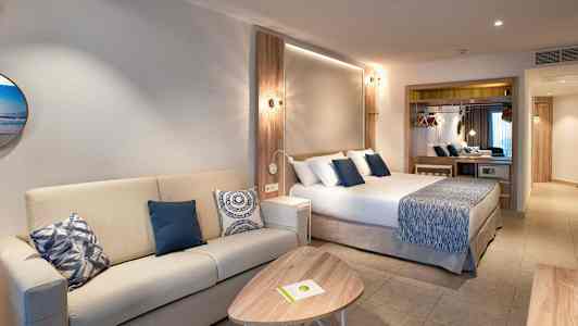 Hotellihuone, jossa on sohva, sohvapöytä ja sinisiä koristeellisia tyynyjä oleva sänky. (Tekoälyn luoma teksti)