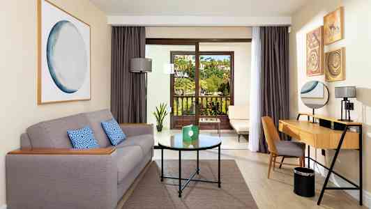 Hotellihuone, jossa on harmaa sohva, kirjoituspöytä ja parveke, josta on näkymä puutarhaan. (Tekoälyn luoma teksti)