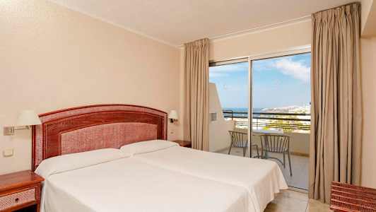 Hotellihuone, jossa on kaksi erillistä sänkyä, puinen pääty ja yöpöydät. Lasipaneelit, jotka tarjoavat näkymät merelle ja parvekekalusteet. (Tekoälyn luoma teksti)