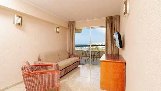 Olohuone, jossa on sohva, nojatuoli, puupöytä ja seinään kiinnitetty televisio. Taustalla on parveke, jolla on tuoleja ja näkymä merelle. (Tekoälyn luoma teksti)