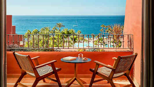 Kaksi tuolia ja pöytä terassilla, jolta on näköala uima-altaalle, palmuihin ja merelle. (Tekoälyn luoma teksti)