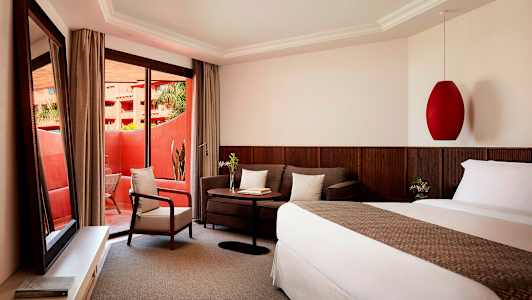 Hotellihuone, jossa on sänky, sohva, pöytä ja nojatuoli. Huoneessa on suuret lasiovet, jotka johtavat parvekkeelle. (Tekoälyn luoma teksti)