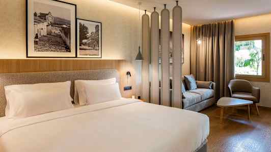 Hotellihuone, jossa on parivuode, istuinryhmä, jossa sohva ja nojatuoli, sekä koristeellisia yksityiskohtia, kuten valokuvia seinällä. (Tekoälyn luoma teksti)