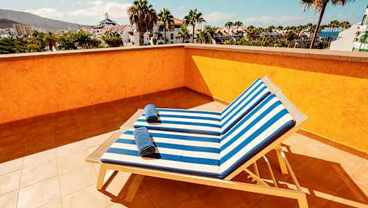 Näkymä aurinkoterassilta, jossa kaksi aurinkotuolia sinivalkoraidallisilla istuinpehmusteilla ja pyyhkeillä, ympäröitynä palmuilla ja rakennuksilla. (Tekoälyn luoma teksti)