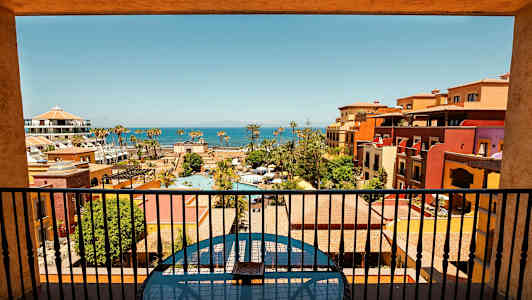 Näkymä parvekkeelta lomakeskukseen, jossa on uima-altaita, värikkäitä rakennuksia ja merimaisema taustalla. (Tekoälyn luoma teksti)