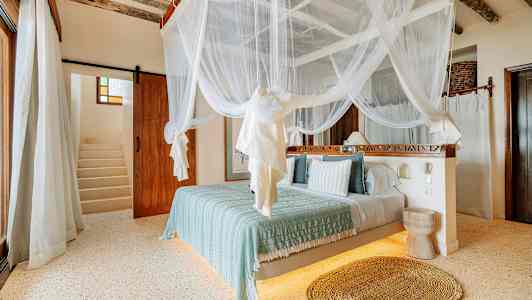 Makuuhuone, jossa on katosvuode valkoisella hyttysverkolla, vaaleat seinät ja puulattia. Taustalla näkyy portaat. (Tekoälyn luoma teksti)