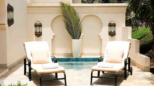 Kaksi aurinkotuolia tyynyillä ja tyynyillä seisoo altaan vieressä vaalean seinän edessä, jossa on koristeelliset lovet ja vihreitä kasveja. (Tekoälyn luoma teksti)