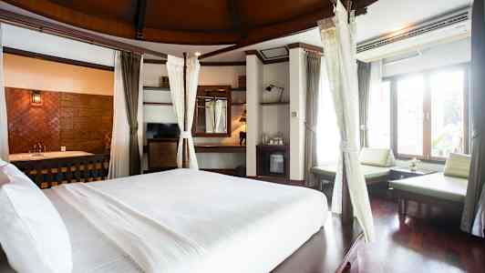 Tilava hotellihuone, jossa on suuri valkoisilla vuodevaatteilla varustettu sänky. Huoneessa on isot ikkunat, jotka päästävät valoa sisään, istuinalue ja puuyksityiskohtia. (Tekoälyn luoma teksti)