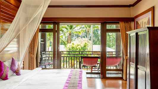 Hotellihuone, jossa on sänky, vaaleanpunaiset tyynyt ja hyttysverkko, lasiovet johtavat parvekkeelle, josta on näkymä palmuihin ja vehreyteen. (Tekoälyn luoma teksti)