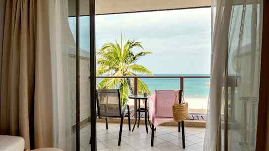 Näkymä hotellihuoneesta parvekkeelle, jossa on kaksi tuolia ja pöytä sekä taustalla ranta ja meri. (Tekoälyn luoma teksti)