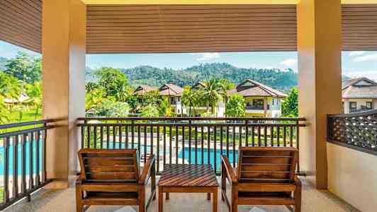 Terassi, jossa on kaksi puista tuolia ja pöytä, joilta on näkymä vehreään puutarhaan, uima-altaaseen ja kerrostaloihin. (Tekoälyn luoma teksti)