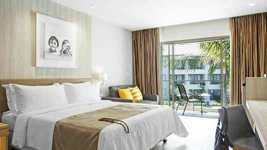 Hotellihuone, jossa on vuode, värikkäillä tyynyillä varustettu sohva sekä pöytä ja tuoli ikkunan ääressä, josta avautuu näkymä parvekkeen ja puutarhan yli. (Tekoälyn luoma teksti)