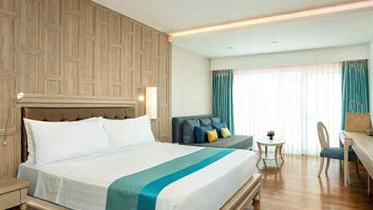 Hotellihuone, jossa on parivuode, sohva tyynyineen, pöytä ja työtuoli. Huoneessa on puupaneloituja seiniä ja lattia sekä suuret ikkunat verhoineen. (Tekoälyn luoma teksti)