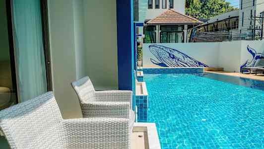 Kaksi valkoista tuolia sijaitsee terassilla uima-altaan vieressä. Altaan viereisellä seinällä on sinisiä maalauksia valaista. (Tekoälyn luoma teksti)