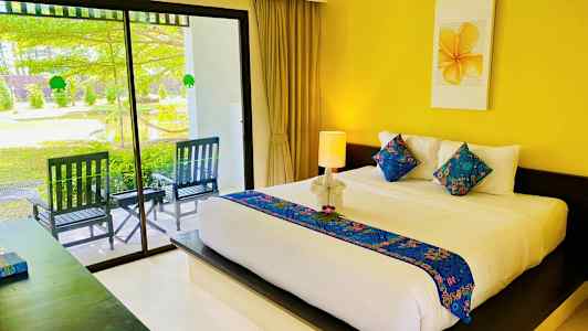 Hotellihuone, jossa on suuri sänky koristeltu värikkäillä tyynyillä ja vaalealla päiväpeitolla. Sänky sijaitsee keltaisen seinän edessä, jonka yläpuolella on maalaus. Lasiovet johtavat ulkoterassille, jossa on puutarha-alue. (Tekoälyn luoma teksti)