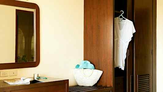 Kuva huoneen sisustuksesta, jossa on peili, lipasto lehden ja tarvikkeiden kanssa sekä avoin vaatekaappi, jossa roikkuu valkoinen vaate. (Tekoälyn luoma teksti)