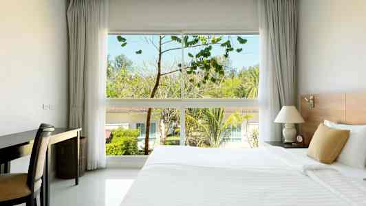 Hotellihuone, jossa on suuri sänky, työpöytä ja ikkuna, josta näkyy puita ja rakennuksia ulkona. (Tekoälyn luoma teksti)