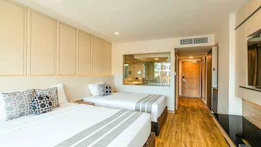 Hotellihuone, jossa on kaksi leveää vuodetta valkoisilla vuodevaatteilla ja koristetyynyillä. Lattia on puinen, ja taustalla on lasiseinä, josta näkyy kylpyhuoneeseen. (Tekoälyn luoma teksti)