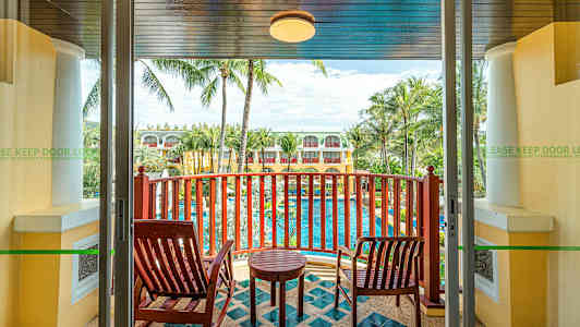 Näkymä parvekkeelta, jolla on kaksi puutuolia ja pieni pöytä, näköala uima-altaalle, jota ympäröivät palmut ja rakennukset. (Tekoälyn luoma teksti)