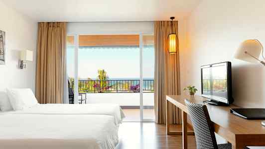 Hotellihuone, jossa on kaksi sänkyä, kirjoituspöytä ja näkymä parvekkeelle, taustalla meri. (Tekoälyn luoma teksti)