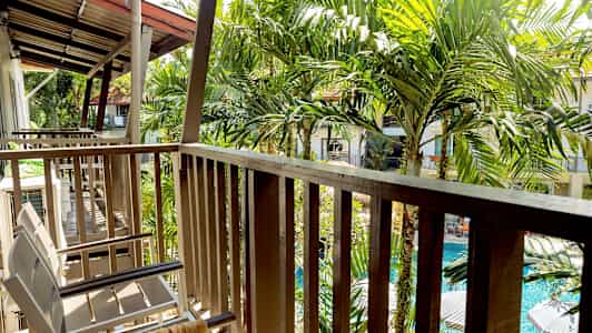 Parveke, jolla on tuoleja ja kaide, ympärillä vihreitä palmuja ja näkymä uima-altaalle. (Tekoälyn luoma teksti)