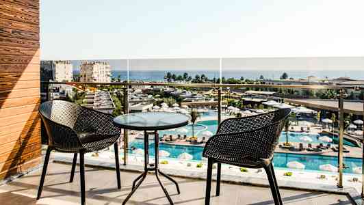 Parveke, jossa kaksi tuolia ja lasipöytä, näkymä uima-allasalueelle ja merelle. (Tekoälyn luoma teksti)