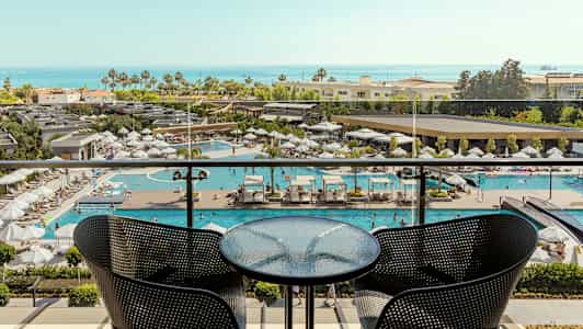 Näkymä parvekkeelta, jossa on pöytä ja kaksi tuolia, näkymä uima-altaalle ja taustalla meri. (Tekoälyn luoma teksti)