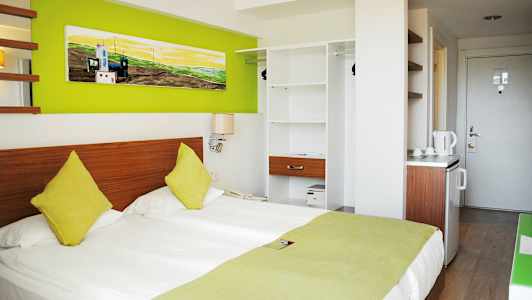 Hotellihuone, jossa on parivuode, värikkäitä tyynyjä, vihreitä sisustuselementtejä ja avoin vaatekaappi. (Tekoälyn luoma teksti)