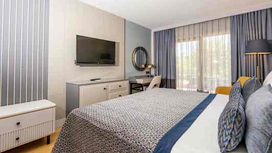 Hotellihuone, jossa moderni sisustus, mukaan lukien sinisillä yksityiskohdilla varustettu sänky, työpöytä, televisio ja suuret ikkunat verhoilla. (Tekoälyn luoma teksti)