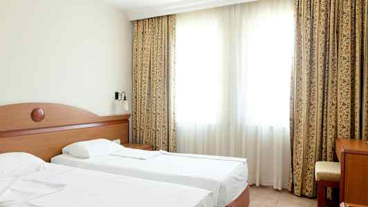 Hotellihuone, jossa on kaksi yhden hengen sänkyä, pöytä ja tuoli suuren ikkunan edessä, sekä verhot. (Tekoälyn luoma teksti)