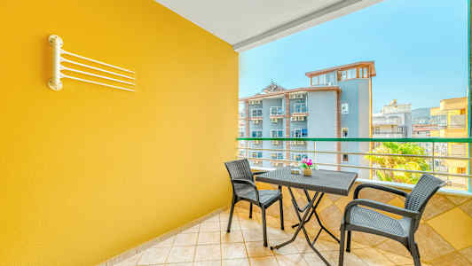 Parveke, jossa on keltainen seinä, pöytä ja kaksi tuolia sekä näkymä muihin rakennuksiin. (Tekoälyn luoma teksti)