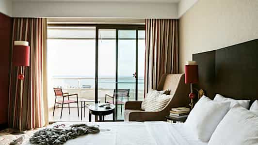 Hotellihuone, jossa on iso sänky, nojatuoli ja parveke merinäköalalla. (Tekoälyn luoma teksti)