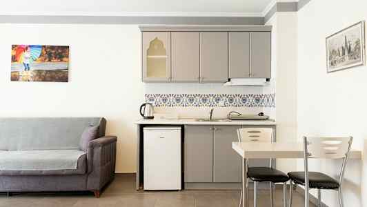 Huone, jossa harmaa sohva, keittiönurkkaus, valkoinen jääkaappi, harmaat kaapit ja värikkäät kaakelit. Vieressä ruokailutila pöydällä ja tuoleilla. (Tekoälyn luoma teksti)