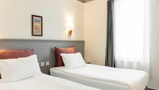 Kaksi sänkyä, joissa on valkoiset lakanat ja punaiset tyynyt, ovat huoneessa, jolla on vaaleat seinät ja seinälamppu. Oikealla on ikkuna valkoisilla verhoilla. (Tekoälyn luoma teksti)