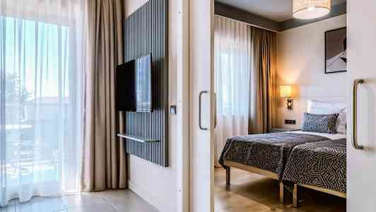 Moderni hotellihuone, jossa on kaksi vuodetta, seinälle asennettu taulu-tv ja suuret ikkunat verhoilla. (Tekoälyn luoma teksti)