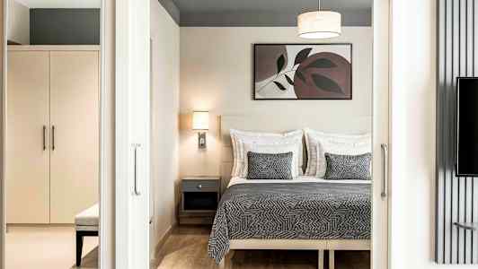 Makuuhuone, jossa on parivuode, harmaa peitto ja yhteensopivat tyynyt, seinälamppu ja sängyn yläpuolella oleva taulu. Vaatekaappi ja penkki näkyvät etualalla. (Tekoälyn luoma teksti)