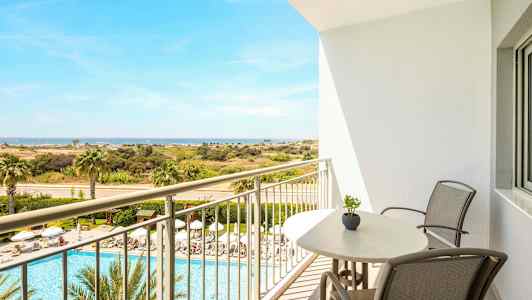 Näköala parvekkeelta, jossa on tuolit ja pöytä, uima-allasalueelle, jossa on aurinkotuoleja ja palmuja, ja edelleen avaraan maisemaan ja merelle. (Tekoälyn luoma teksti)