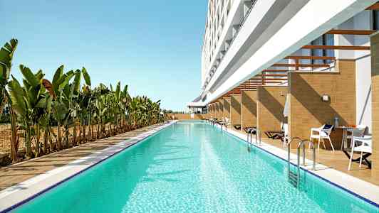 Pitkä uima-allas, jossa kirkas sininen vesi, rakennuksen vieressä, jossa on yksityisiä terasseja ja aurinkotuoleja, ympärillä banaanipuita. (Tekoälyn luoma teksti)