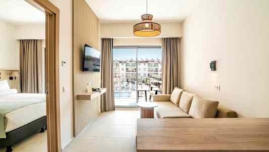 Hotellihuone, jossa on istuinryhmä, sänky ja televisio, sekä parveke, josta on näkymä allasalueelle. (Tekoälyn luoma teksti)