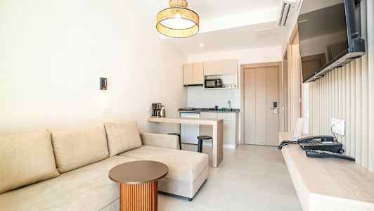 Hotellihuone, jossa on beige sohva, pieni pöytä, keittiö ja seinälle asennettu televisio. (Tekoälyn luoma teksti)