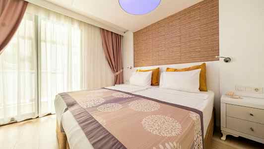 Hotellihuone, jossa parivuode, koristeltu kuvioidulla päiväpeitolla ja tyynyillä. Ikkuna läpinäkyvillä verhoilla ja violetit verhot taustalla. (Tekoälyn luoma teksti)