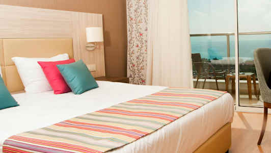 Hotellihuone, jossa on parivuode, värikkäitä tyynyjä ja näkymä merelle parvekkeelta. (Tekoälyn luoma teksti)