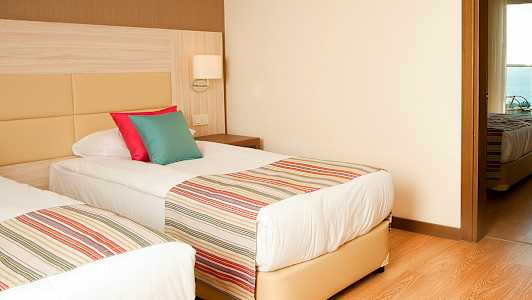 Kaksi yhden hengen vuodetta raidallisilla päiväpeitteillä ja värikkäillä tyynyillä, huoneessa, jossa on puulattia ja vaaleat seinät. (Tekoälyn luoma teksti)