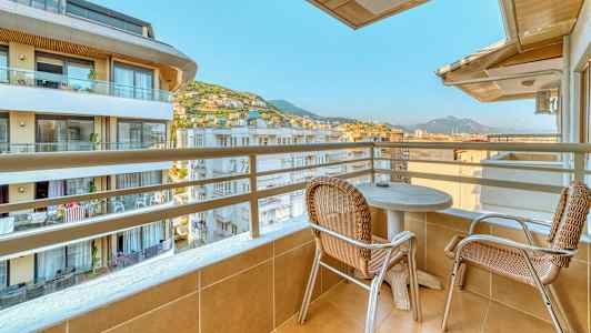 Parveke, jolla on kaksi tuolia ja pöytä, näkymä rakennuksiin ja vuoristoihin. (Tekoälyn luoma teksti)