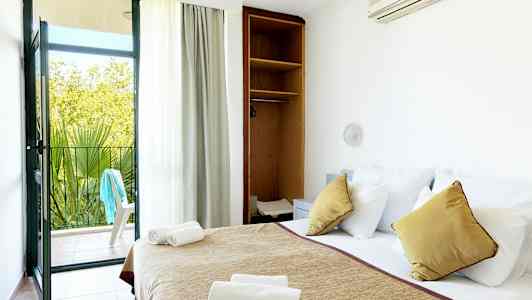 Hotellihuone, jossa on pedattu sänky, avoin kaappi ja parvekkeen ovi, joka johtaa ulkopatiolle. (Tekoälyn luoma teksti)