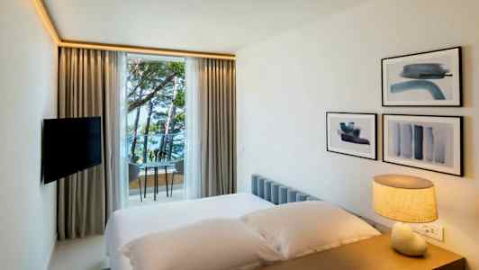 Hotellihuone, jossa on sänky, pöydällä oleva lamppu, seinäkoristeita ja ikkuna, joka avautuu merelle ja puille. (Tekoälyn luoma teksti)