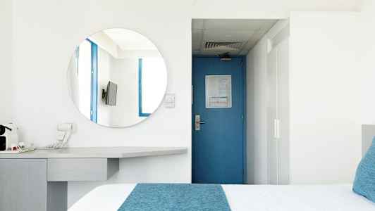 Hotellihuone, jossa sininen ovi, pyöreä peili, televisio ja sinisiä yksityiskohtia sisältävä sänky. (Tekoälyn luoma teksti)